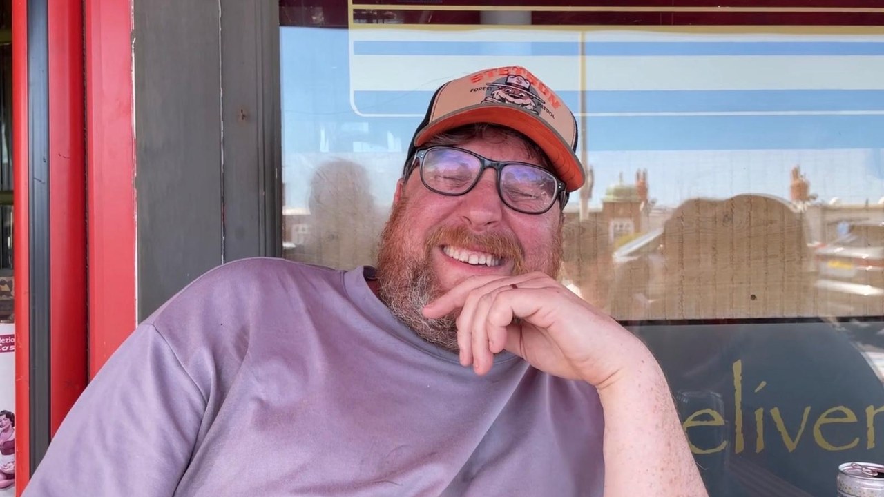 Ben Rymer, smiling, sitting outside a shopfront on a sunny day.